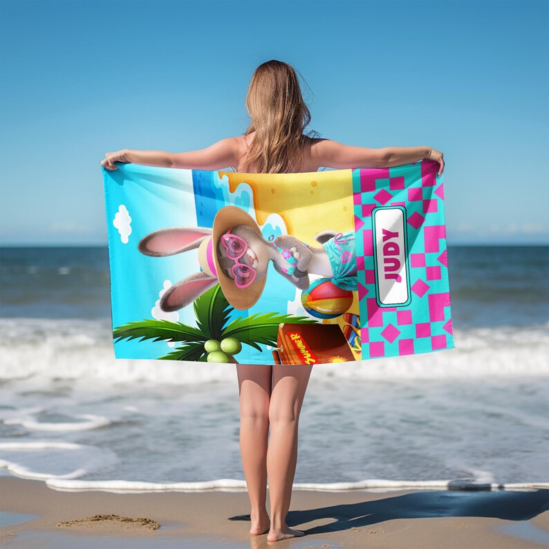 Może przedstawiać: Prostokątny ręcznik plażowy z rysunkowym kr&oacute;likiem w słomkowym kapeluszu i r&oacute;żowych okularach przeciwsłonecznych w kształcie serc. Wz&oacute;r przedstawia plażę z palmą, piłką plażową oraz r&oacute;żowo-niebieski geometryczny motyw z imieniem JUDY.