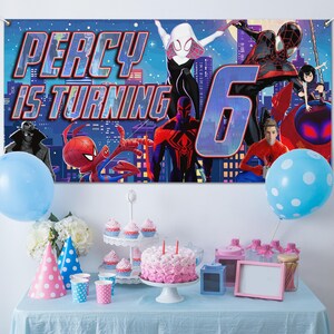 May include: A rectangular party banner featuring Spider-Man characters against a city skyline with the text PERCY IS TURNING 6. Below, a table holds a pink frosted cake, cupcakes on a tiered stand, party hats, and blue polka dot balloons.