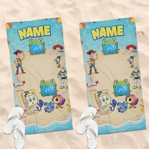 May include: Two beach towels with a cartoon beach scene featuring characters. The towels have a blue border with the word "NAME" in yellow. The beach scene includes cartoon characters, a frog with "Hi!" on a sign, and white flip-flops.