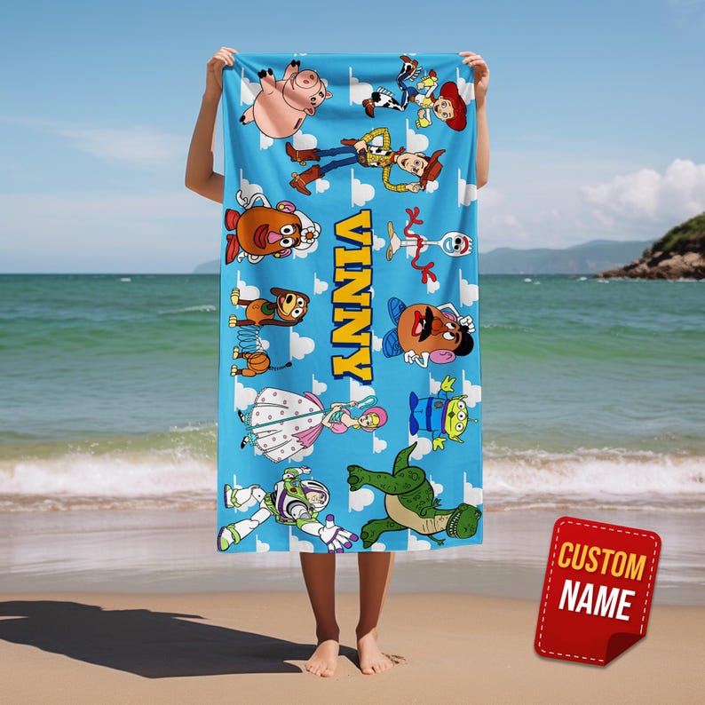 May include: A blue beach towel featuring cartoon characters from the Toy Story franchise. The towel has the name "VINNY" in yellow block letters. The characters include Woody, Buzz Lightyear, and others. A red tag with "CUSTOM NAME" is in the bottom right corner.