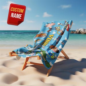 May include: A blue beach towel featuring cartoon characters, draped over a wooden beach chair on a sandy beach. The towel has the name "VINNY" printed on it. A red tag with the text "CUSTOM NAME" is in the upper left corner.