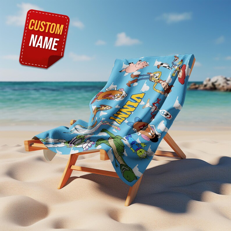 May include: A blue beach towel featuring cartoon characters, draped over a wooden beach chair on a sandy beach. The towel has the name "VINNY" printed on it. A red tag with the text "CUSTOM NAME" is in the upper left corner.