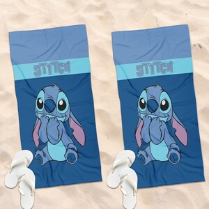 May include: Two rectangular blue beach towels featuring a graphic of the character Stitch with the text STITCH printed in light blue. Each towel is paired with a pair of white flip-flops, displaying a consistent design across both items.
