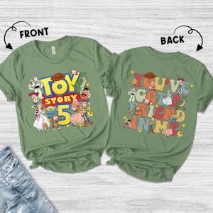 K&ouml;nnte beinhalten: Zwei olivgr&uuml;ne T-Shirts mit den Aufdrucken "Toy Story 5" und "You've Got a Friend in Me". Das vordere Shirt zeigt verschiedene Toy Story-Charaktere. Das hintere Shirt hat die gleichen Charaktere und den gleichen Text.