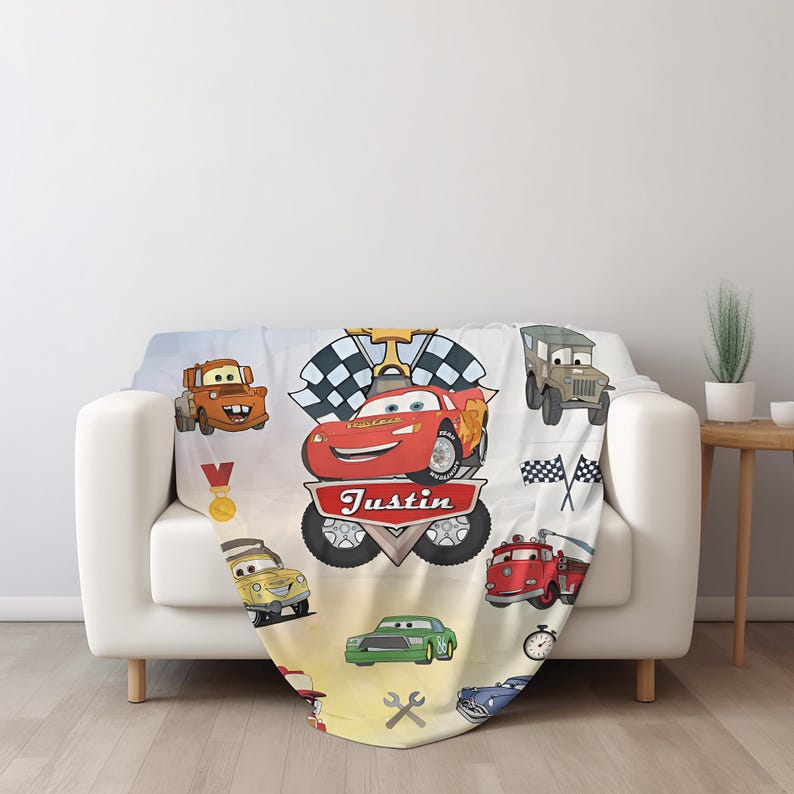 May include: A white fleece blanket featuring a colorful cartoon race car theme with various vehicles, a checkered flag, a gold medal, and a stopwatch. The name Justin is printed in white letters on a red banner centered below a red race car.