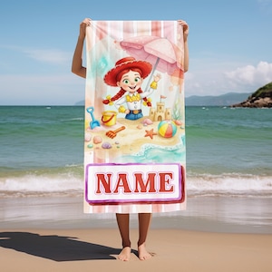 Puede incluir: Una toalla de playa rectangular con un personaje de dibujos animados de pelo rojo trenzado y traje de vaquera, bajo una sombrilla rosa en una playa de arena con un castillo de arena, un cubo, una pala y una pelota. La palabra NAME est&aacute; en rojo.