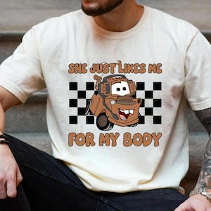Puede incluir: Una camiseta de manga corta color crema con un gr&aacute;fico de una gr&uacute;a marr&oacute;n que lleva auriculares. Detr&aacute;s del veh&iacute;culo hay un patr&oacute;n de cuadros blancos y negros. El texto dice: SHE JUST LIKES ME FOR MY BODY en letras de color marr&oacute;n.