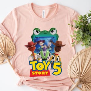 May include: A light pink t-shirt featuring a graphic of characters from the Toy Story movie franchise. The graphic includes the text "TOY 5 STORY" in yellow and red. The shirt has a round neckline and short sleeves.