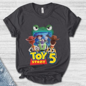 May include: Dark gray t-shirt featuring a colorful graphic from the movie Toy Story 5. The design includes characters Woody, Buzz Lightyear, and Jessie, with the words "TOY STORY 5" in yellow and red. The shirt is made of a soft material.