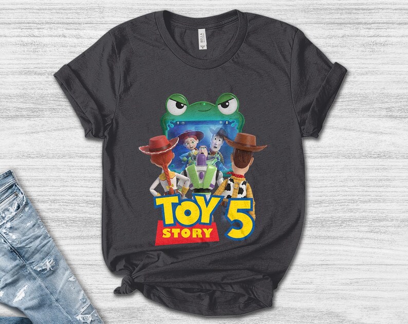 May include: Dark gray t-shirt featuring a colorful graphic from the movie Toy Story 5. The design includes characters Woody, Buzz Lightyear, and Jessie, with the words "TOY STORY 5" in yellow and red. The shirt is made of a soft material.