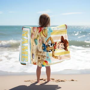 Op de afbeelding: Een rechthoekige strandhanddoek met een cartoon cowboyfiguur, een zandkasteel, een gele emmer en een blauwe schep op een zandstrandachtergrond. De handdoek heeft een verticale blauwe rand met het woord NAME in grote gele letters gedrukt.