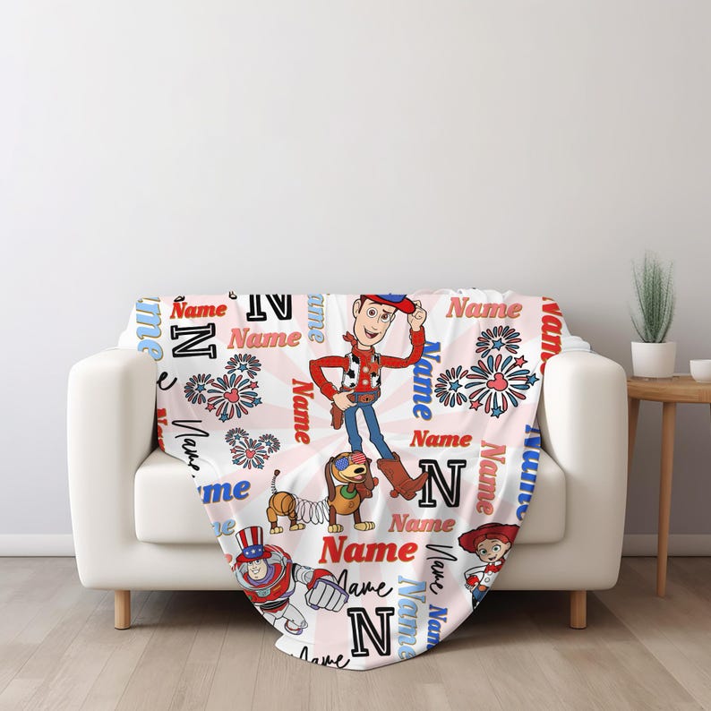 Op de afbeelding: Een witte deken met stripfiguren en het woord "Name" in rood en blauw. De deken heeft een herhalend patroon van sterren, harten en de letter "N". De deken is over een witte bank gedrapeerd.