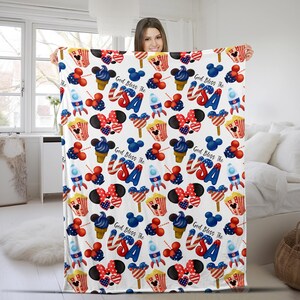 Puede incluir: Una manta blanca con un patr&oacute;n repetido de iconos patri&oacute;ticos estadounidenses. El dise&ntilde;o incluye conos de helado rojos, blancos y azules, orejas de Mickey Mouse, cohetes, palomitas de ma&iacute;z y el texto "God Bless The USA".