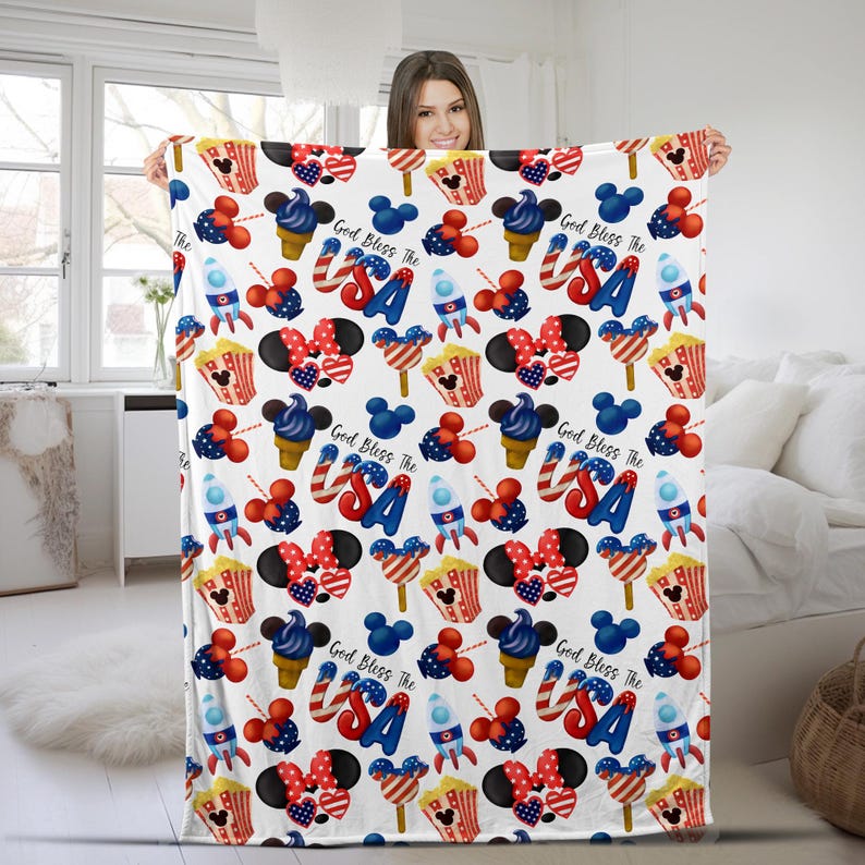 Puede incluir: Una manta blanca con un patr&oacute;n repetido de iconos patri&oacute;ticos estadounidenses. El dise&ntilde;o incluye conos de helado rojos, blancos y azules, orejas de Mickey Mouse, cohetes, palomitas de ma&iacute;z y el texto "God Bless The USA".