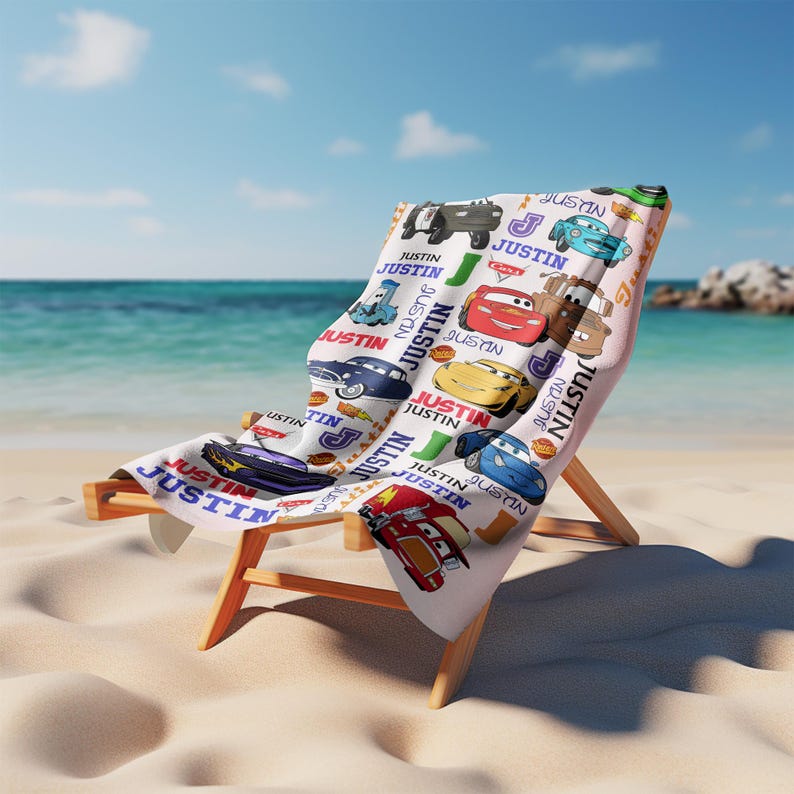 May include: A beach chair with a personalized towel featuring cartoon cars and the name "JUSTIN" in a repeating pattern. The towel is draped over the wooden chair on a sandy beach with the ocean in the background.