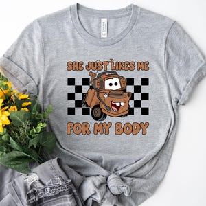 Puede incluir: Una camiseta gris jaspeado con un gr&aacute;fico de una gr&uacute;a marr&oacute;n con auriculares, centrado sobre un patr&oacute;n de cuadros blancos y negros. El texto encima y debajo del personaje dice "SHE JUST LIKES ME FOR MY BODY" en color marr&oacute;n.