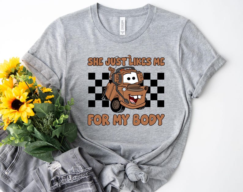 Puede incluir: Una camiseta gris jaspeado con un gr&aacute;fico de una gr&uacute;a marr&oacute;n con auriculares, centrado sobre un patr&oacute;n de cuadros blancos y negros. El texto encima y debajo del personaje dice "SHE JUST LIKES ME FOR MY BODY" en color marr&oacute;n.