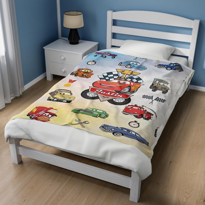 May include: A white bed blanket featuring a colorful cartoon car print with various vehicles, checkered flags, a gold trophy, and a red banner with the name Justin printed in white. The design includes multiple race cars, a fire truck, and tools.