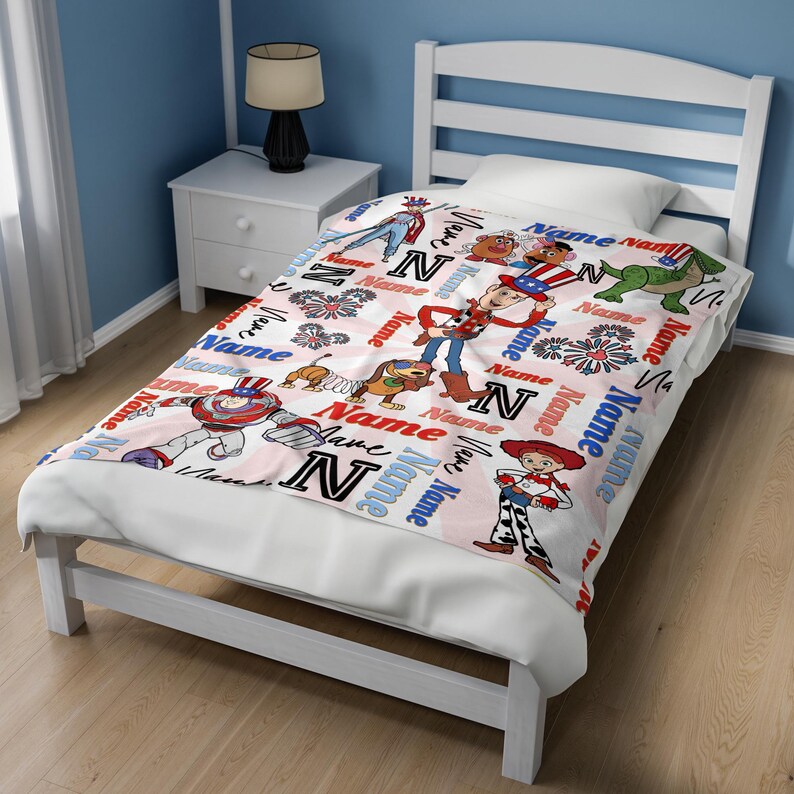 Op de afbeelding: Een wit en roze deken met stripfiguren en het woord "Name" in rood en blauw. De deken heeft een herhalend patroon van karakters en de letter "N".