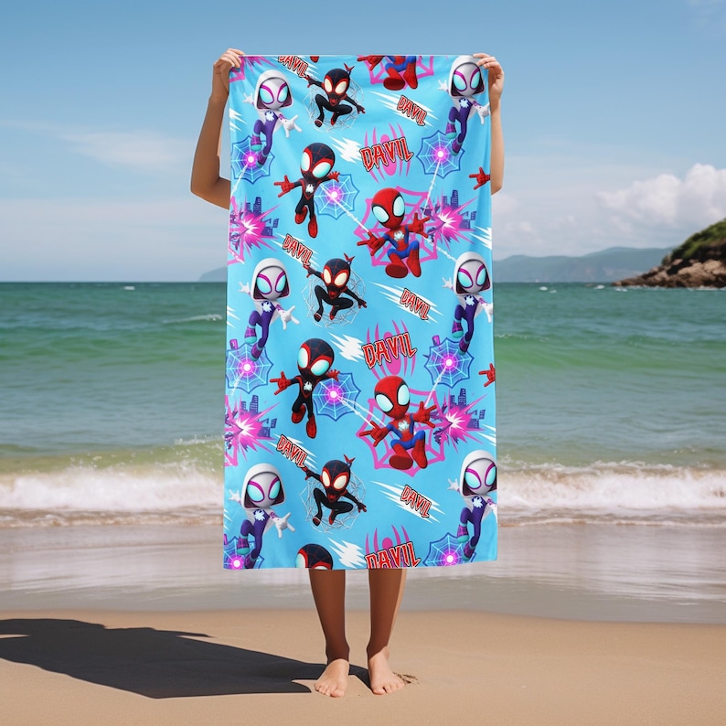 Puede incluir: Una toalla de playa rectangular de color azul claro con un patr&oacute;n repetitivo de superh&eacute;roes de dibujos animados en trajes rojos, negros y blancos, telara&ntilde;as rosas y el nombre DAVIL impreso en letras rojas. La toalla muestra varias poses.