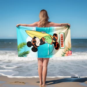 K&ouml;nnte beinhalten: Ein rechteckiges Strandtuch mit einer Cartoon-Illustration von Mickey Mouse, der eine orangefarbene Sonnenbrille und eine gr&uuml;ne Shorts tr&auml;gt und ein gelbes Surfbrett h&auml;lt. Der Hintergrund zeigt einen Strand mit Palmen und dem Text MICKEY.
