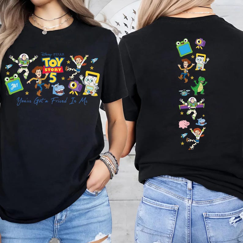 May include: A black t-shirt featuring Toy Story 5 graphics. The front displays the Toy Story 5 logo, characters including Woody, Buzz Lightyear, and Jessie, and the text You've Got a Friend In Me. The back shows a vertical column of characters.