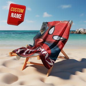 May include: A red and blue Spider-Man beach towel draped over a wooden beach chair on a sandy beach. The towel features a close-up of Spider-Man's face and the text "CUSTOM NAME" on a red sticker.
