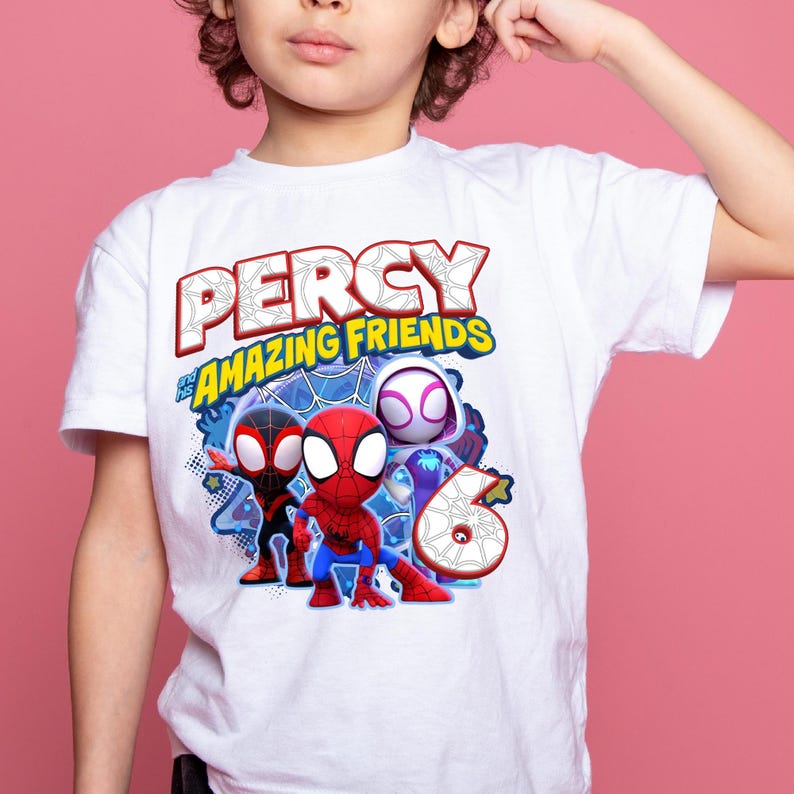 May include: A white short-sleeved t-shirt featuring a graphic print with the text PERCY and his AMAZING FRIENDS. The design includes three superhero characters in red, black, and white costumes, along with a large number six with a spiderweb pattern.