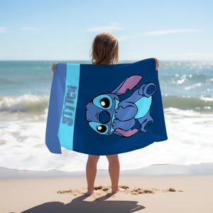 May include: A rectangular beach towel featuring a dark blue background with a large graphic of the character Stitch. The towel includes a light blue vertical stripe on the left side with the word STITCH printed in a bold, blue, outlined font.