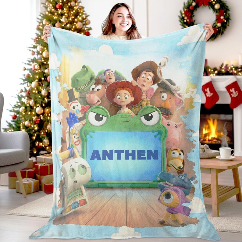 May include: A light blue fleece blanket featuring a group of animated characters from Toy Story surrounding a large green frog-shaped frame with the name ANTHEN printed in bold blue capital letters in the center against a wooden floor background.