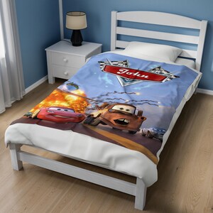 May include: A blue bed blanket featuring a graphic print of the characters Lightning McQueen and Mater from the movie Cars. The design includes a red banner with the name John written in white script, checkered flags, and an explosion effect.