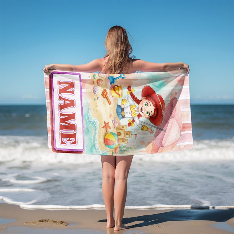 Puede incluir: Una toalla de playa rectangular con un personaje de vaquera de dibujos animados con trenzas rojas, un castillo de arena, una pelota de playa y una pala. El dise&ntilde;o incluye un recuadro vertical con borde morado y la palabra NAME en letras rojas.