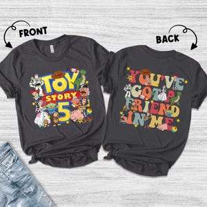 K&ouml;nnte beinhalten: Zwei dunkelgraue T-Shirts mit den Aufdrucken "TOY STORY 5" und "YOU'VE GOT A FRIEND IN ME". Das vordere Shirt zeigt verschiedene Zeichentrickfiguren. Das hintere Shirt hat die gleichen Figuren und den gleichen Text.