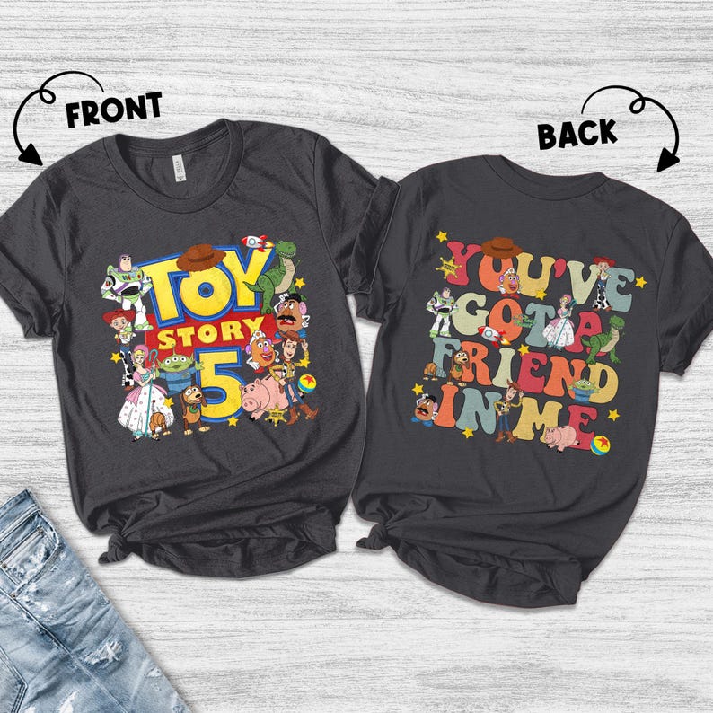 K&ouml;nnte beinhalten: Zwei dunkelgraue T-Shirts mit den Aufdrucken "TOY STORY 5" und "YOU'VE GOT A FRIEND IN ME". Das vordere Shirt zeigt verschiedene Zeichentrickfiguren. Das hintere Shirt hat die gleichen Figuren und den gleichen Text.