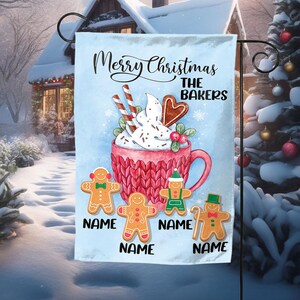 May include: A personalized Christmas garden flag with a festive design. It features a mug of hot cocoa with whipped cream, gingerbread cookies, and the text "Merry Christmas The Bakers." There are spaces for names.