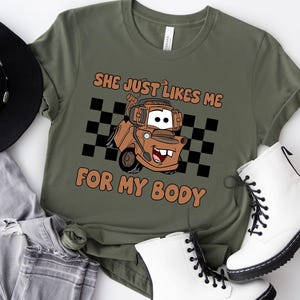 Puede incluir: Una camiseta verde oliva con un gr&aacute;fico de una gr&uacute;a de dibujos animados marr&oacute;n con auriculares, situada frente a un patr&oacute;n de cuadros blancos y negros. El texto "SHE JUST LIKES ME FOR MY BODY" est&aacute; impreso en naranja sobre y bajo el cami&oacute;n.