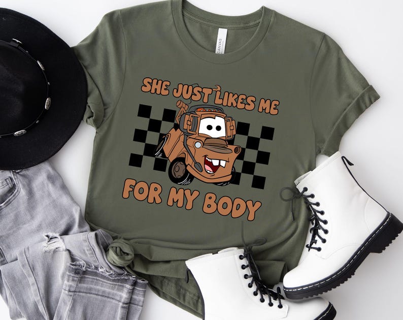 Puede incluir: Una camiseta verde oliva con un gr&aacute;fico de una gr&uacute;a de dibujos animados marr&oacute;n con auriculares, situada frente a un patr&oacute;n de cuadros blancos y negros. El texto "SHE JUST LIKES ME FOR MY BODY" est&aacute; impreso en naranja sobre y bajo el cami&oacute;n.