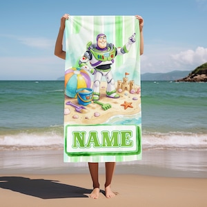 May include: A rectangular beach towel featuring a cartoon space ranger character standing on a sandy beach with a beach ball, a blue bucket, a sandcastle, and a starfish. The towel has a green and white striped background and a green box with the text NAME.