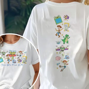 May include: A white t-shirt featuring Toy Story 5 graphics. The front displays the Toy Story 5 logo with characters and the text "You've Got a Friend In Me." The back shows a vertical arrangement of various Toy Story characters including Woody, Buzz Lightyear, Rex, and Jessie.