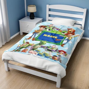May include: A light blue fleece blanket featuring a colorful graphic print of various animated toy characters, including a cowboy, a space ranger, and a dinosaur. The center of the blanket displays a green frame with the yellow text NAME.