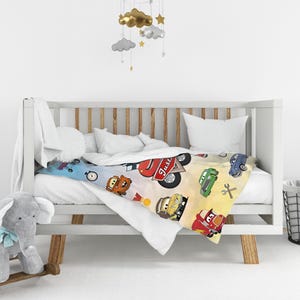 May include: A white crib blanket featuring a colorful pattern of cartoon race cars, a checkered flag, a gold medal, and a wrench. The design includes a red car with the text Just printed in white script on a light blue and yellow background.