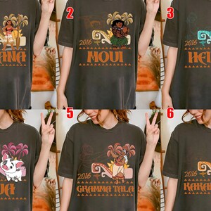 May include: A charcoal gray t-shirt featuring a graphic print of a Moana character, the year 2016, palm trees, and a decorative border. The design includes the character name in bold orange letters at the bottom, with six variations shown.