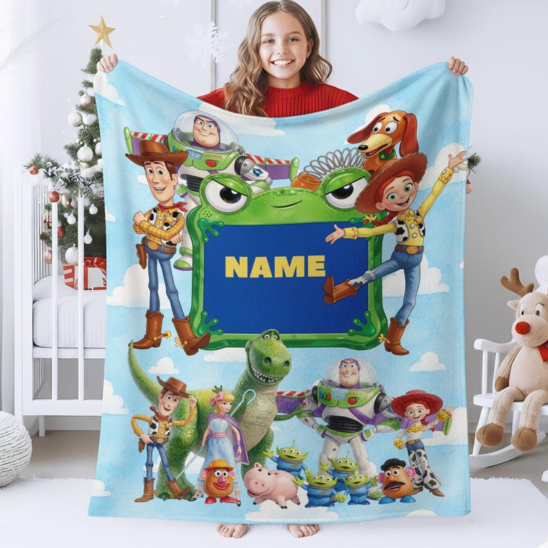 May include: A light blue fleece blanket featuring a colorful print of Toy Story characters including Woody, Buzz Lightyear, Jessie, Rex, and Slinky Dog. A central green frog-shaped frame contains the yellow text NAME against a dark blue background.
