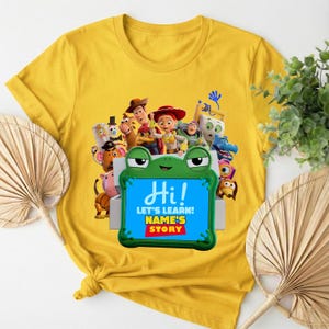 May include: Mustard yellow t-shirt with a vibrant graphic of animated characters. The design includes the text "Hi!" and "LET'S LEARN! NAME'S STORY" on a blue background, framed by a green frog.