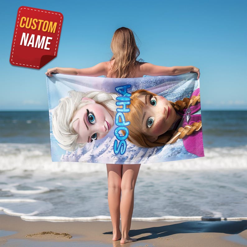 Pode incluir: Uma toalha de praia com Elsa e Anna de Frozen, com o nome "Sophia" em escrita azul. A toalha &eacute; segurada por uma pessoa em uma praia com o oceano ao fundo. Um adesivo vermelho no canto diz "CUSTOM NAME".