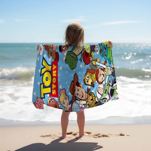 May include: A light blue beach towel featuring characters from the animated film "Toy Story." The towel has a repeating pattern of the characters and the words "Toy Story" in large yellow letters. The towel is held up on a beach with the ocean in the background.