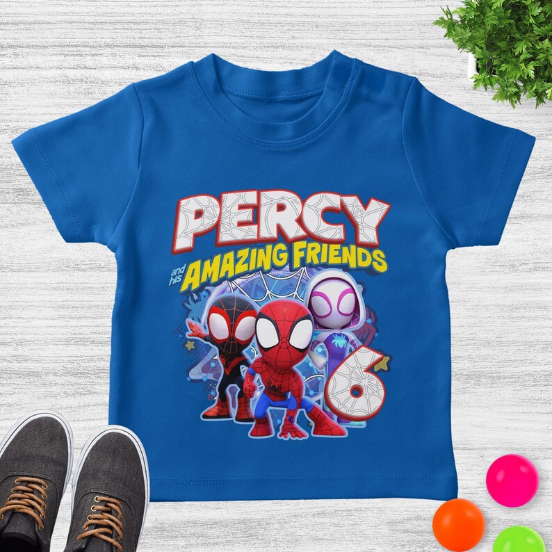 May include: A royal blue short-sleeve t-shirt featuring a graphic print with the text PERCY and his AMAZING FRIENDS. The design includes three cartoon spider characters and a large number 6, all set against a blue spiderweb background pattern.