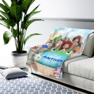 May include: A soft throw blanket featuring a colorful graphic print of various animated toy characters, including a prominent green frog-shaped device with the name ANTHEN printed in bold blue capital letters across its center surface.