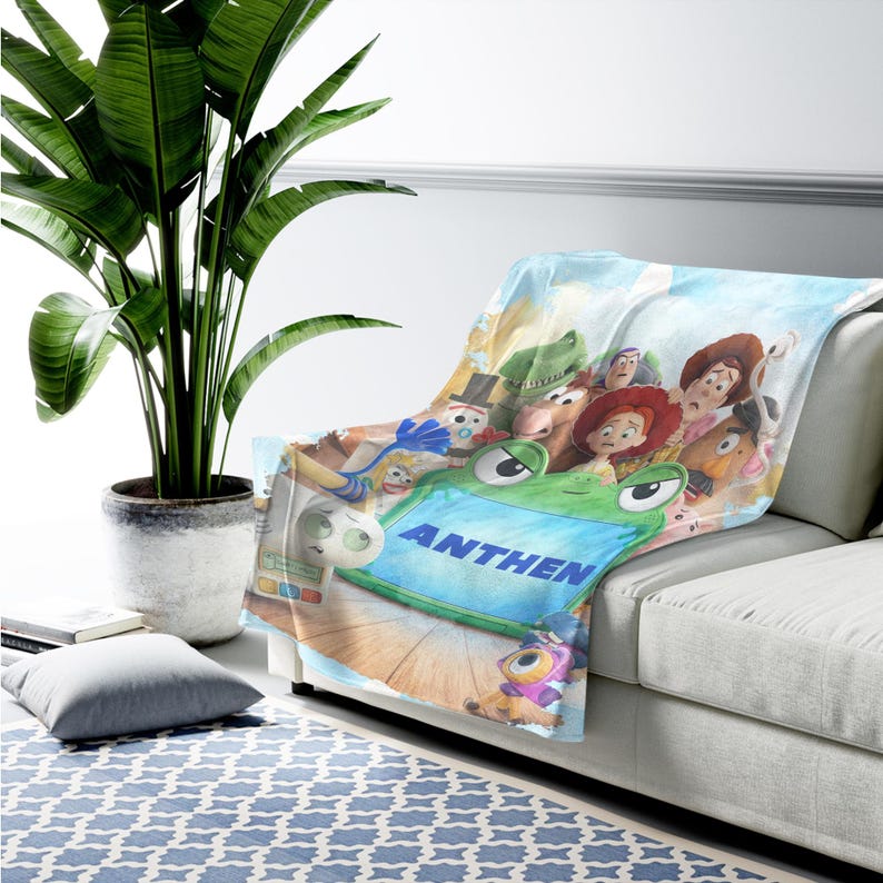 May include: A soft throw blanket featuring a colorful graphic print of various animated toy characters, including a prominent green frog-shaped device with the name ANTHEN printed in bold blue capital letters across its center surface.