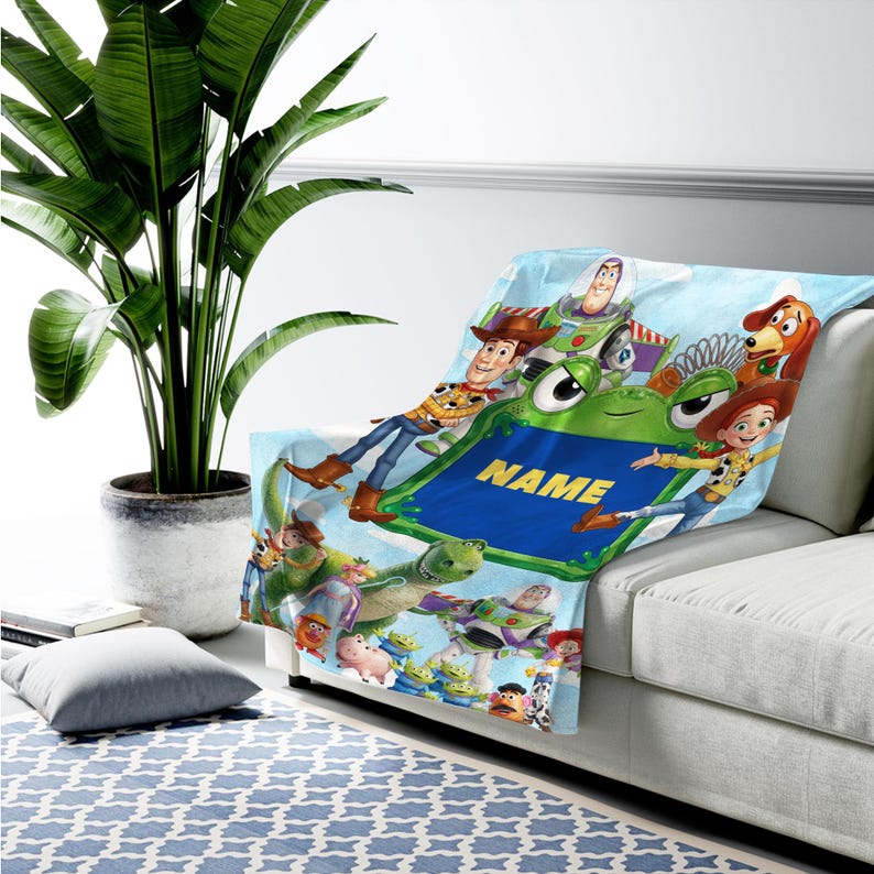 May include: A soft fleece blanket featuring a colorful print of various animated characters from the Toy Story franchise, including Woody, Buzz Lightyear, and Jessie. The center displays a green frame with the yellow text NAME in capital letters.